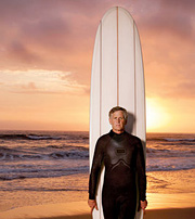 A middle aged man stands in front of his surfboard and the ocean. - Copyright – Stock Photo / Register Mark